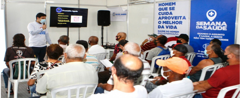 Foto feita numa palestra onde há 15 pessoas sentadas de costas e um palestrante ao fundo de frente para o fotógrafo. É possível ler mensagens de banners fixados na parede da sala, como "Semana da Saúde" e "Quando o assunto é saúde, a gente não tem tempo a perder"