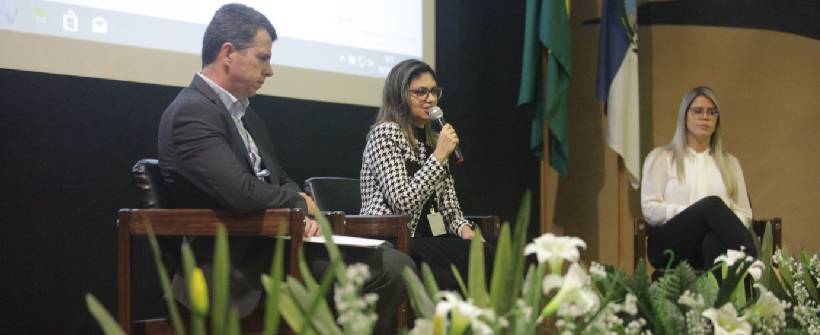 Foto mostra um homem e duas mulehres. Uma delas, no meio, fala ao microfone.