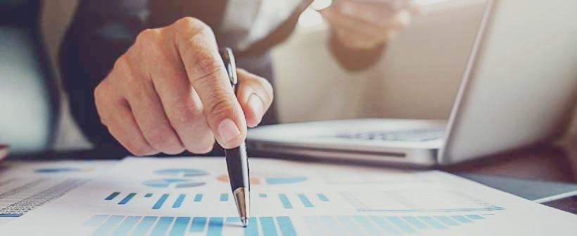 A imagem mostra uma pessoa segurando uma caneta e uma planilha impressa em papel
