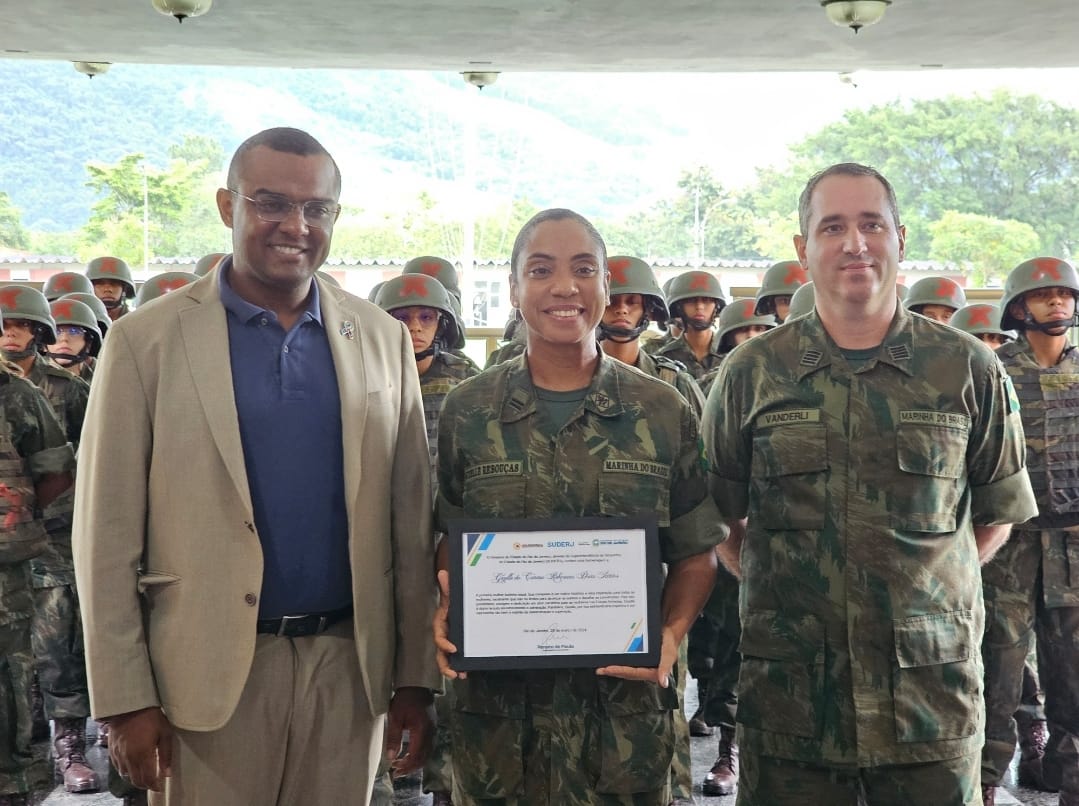 uma mulher fardada com uma placa nas mãos, ao lado de um homem de terno e outro fardado.