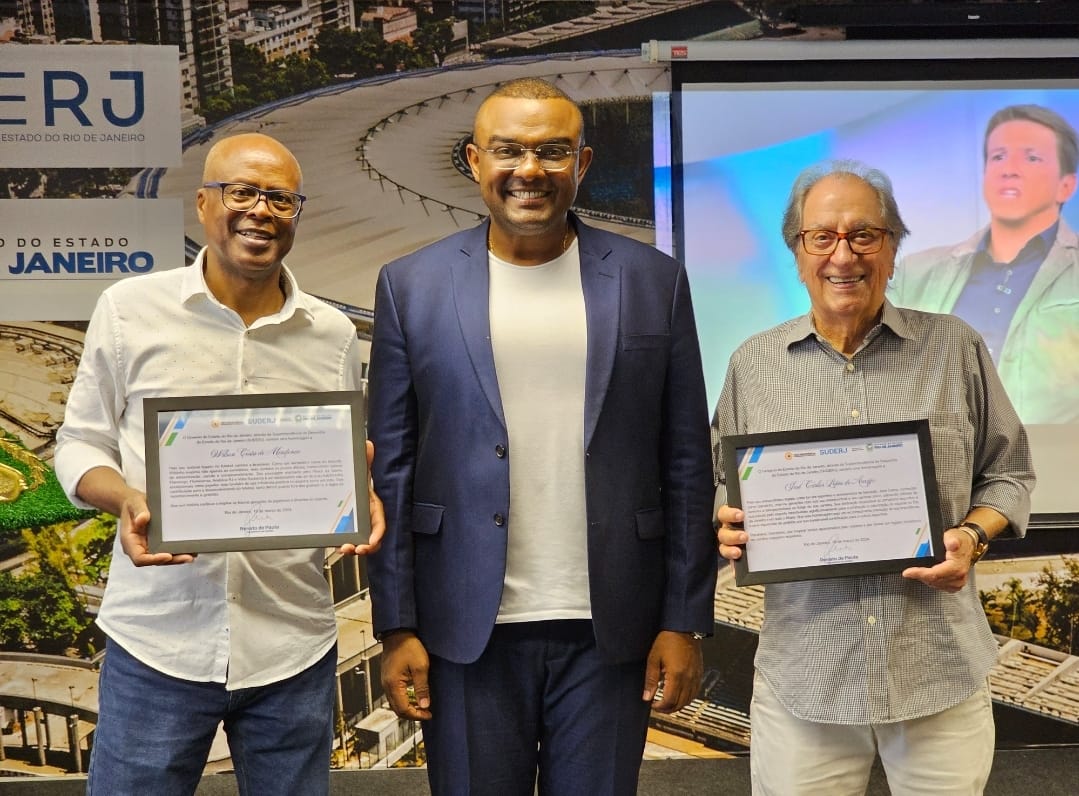 Três homens sorrindo para foto, onde dois estão segurando placas de homenagem.